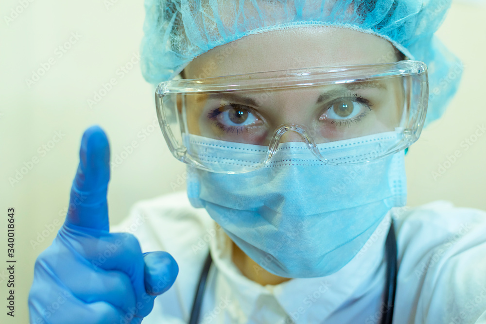 A female doctor in a medical mask, glasses, and gloves sternly hold up her index finger, a sign of advice to be careful. A female doctor in a medical mask, glasses, and gloves sternly hold up her index finger, a sign of advice to be careful.