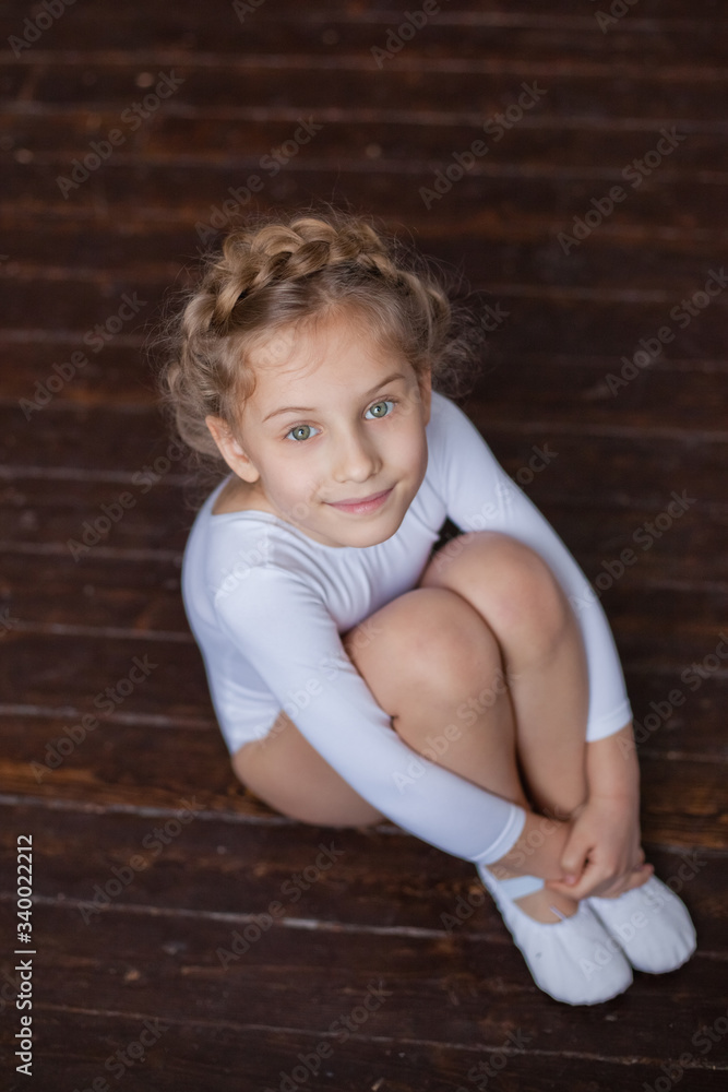 Naklejka premium A charming little girl in a white gymnastic costume is training in rhythmic gymnastics. A little girl is doing exercises on the floor. For a child, the concept