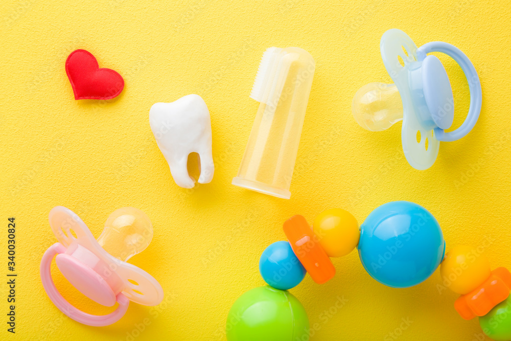 Red heart, soothers, white tooth and transparent silicone fingertip toothbrush and colorful rattle on bright yellow table background. Things for babies teeth cleaning through fun. Close up. Top view.