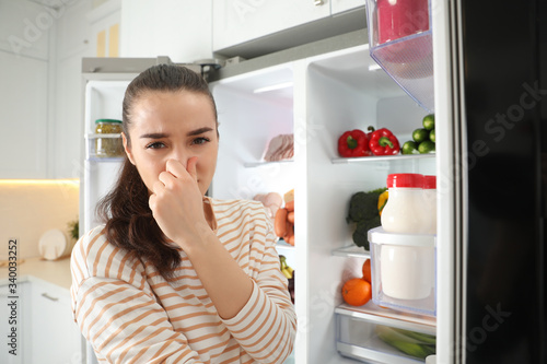 Young woman holding nose cause of bad smell in refrigerator in kitchen