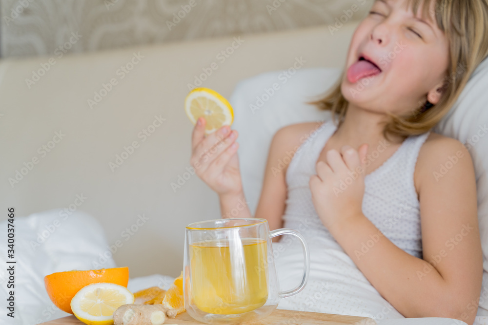 Sick girl in bed tastes lemon slice.Cup of antipyretic drugs for colds,flu.Tea with citrus vitamin C,ginger root,orange.Wooden tray.Home self-treatment.Medical quarantine covid-19 coronavirus therapy Sick girl in bed tastes lemon slice.Cup of antipyretic drugs for colds,flu.Tea with citrus vitamin C,ginger root,orange.Wooden tray.Home self-treatment.Medical quarantine covid-19 coronavirus therapy