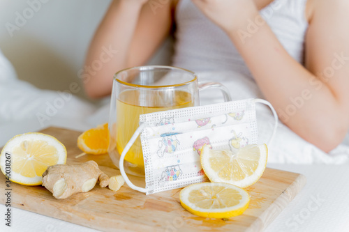 Sick girl in bed tastes orange.Children face mask.Cup of tea with citrus vitamin C,ginger root,lemon.Drugs for cold,flu.Wooden tray.Home self-treatment.Medical quarantine therapy.Covid-19,coronavirus
