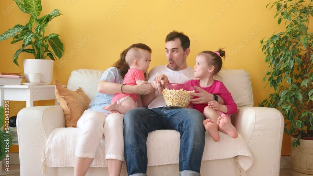Young Family with Two Little Daughters Sitting on the Couch and Watching TV and Eating Popcorn Together in Cozy Home Interior. Young Family with Two Little Daughters Sitting on the Couch and Watching TV and Eating Popcorn Together in Cozy Home Interior.