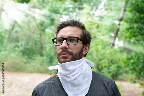 A young bearded man with brown hair and prescription glasses stands in the wilderness wearing a white bandana and a grey and yellow rain jacket looking ahead after just getting rained.