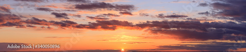 panorama view of colorful sky at sunset with the setting sun and orange yellow purple clouds in front of twilight and the last sunbeams, wide picture  © Maxim Kukurund