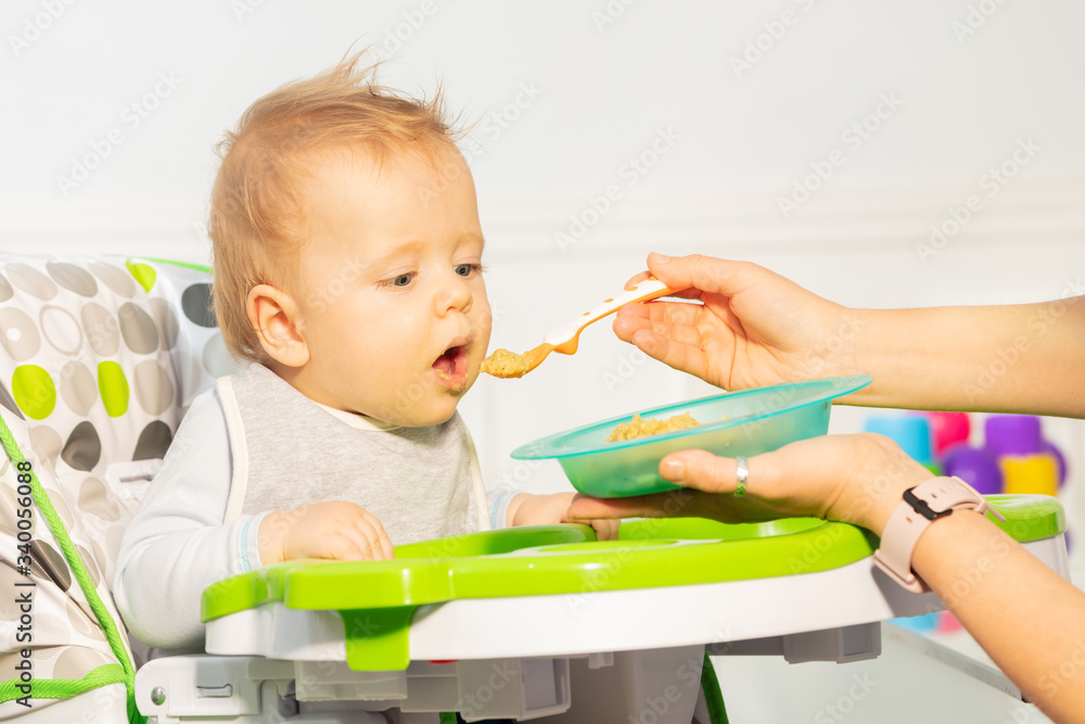 Naklejka premium Little toddler baby boy sit in high chair eager to eat food from spoon proposed by mother