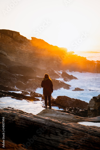 Girl standing on a rock watching the sunset while waves crash against the coastline