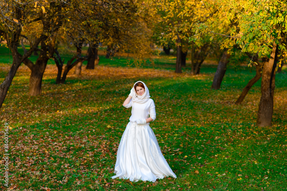 Cute girl in long white wedding dress posing in rural path among autumnal trees in forest in golden hour atmosphere.