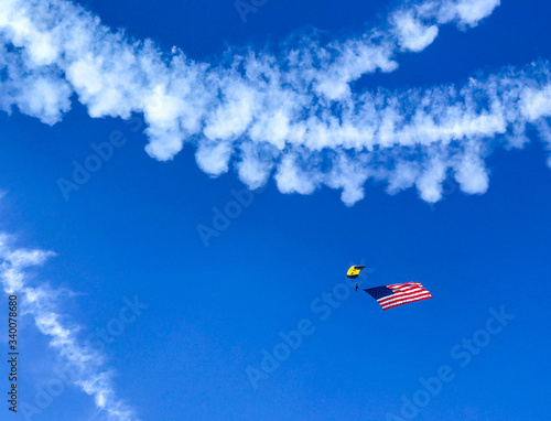 Stunt Skydiver with Parachute and an American Flag against Blue Sky with Smoke Airplane Contrails