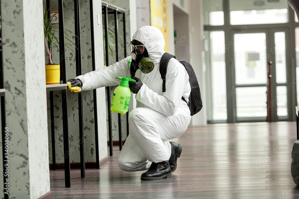 Fototapeta premium A man in protective equipment disinfects with a sprayer in the office. Surface treatment due to coronavirus covid-19 disease. A man in a white suit disinfects the room with a spray gun. Virus pandemic