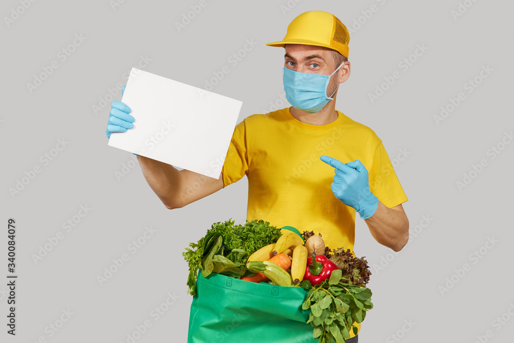 Online shopping. Male courier in yellow uniform, protective mask and gloves with a grocery box with fresh fruits and vegetables holds a White banner for text. Home delivery food during quarantine Online shopping. Male courier in yellow uniform, protective mask and gloves with a grocery box with fresh fruits and vegetables holds a White banner for text. Home delivery food during quarantine