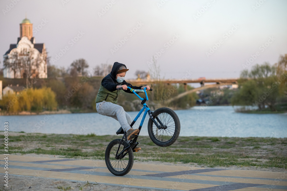 A masked teenager on a blue-black bicycle rides on the rear wheel. River embankment. The church and the bridge in the background. Covid-19.