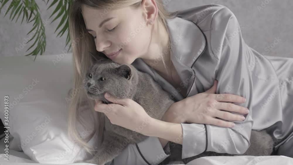 woman in gray pajamas lying on the bed hugs a cat of breed British woman in gray pajamas lying on the bed hugs a cat of breed British