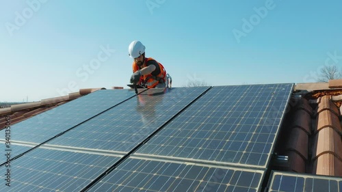 Aerial wide shot of worker with hard hat and safety equipment installing photovoltaic solar panel system installed on home domestic roof top, urban landscape. eco responsible business concept