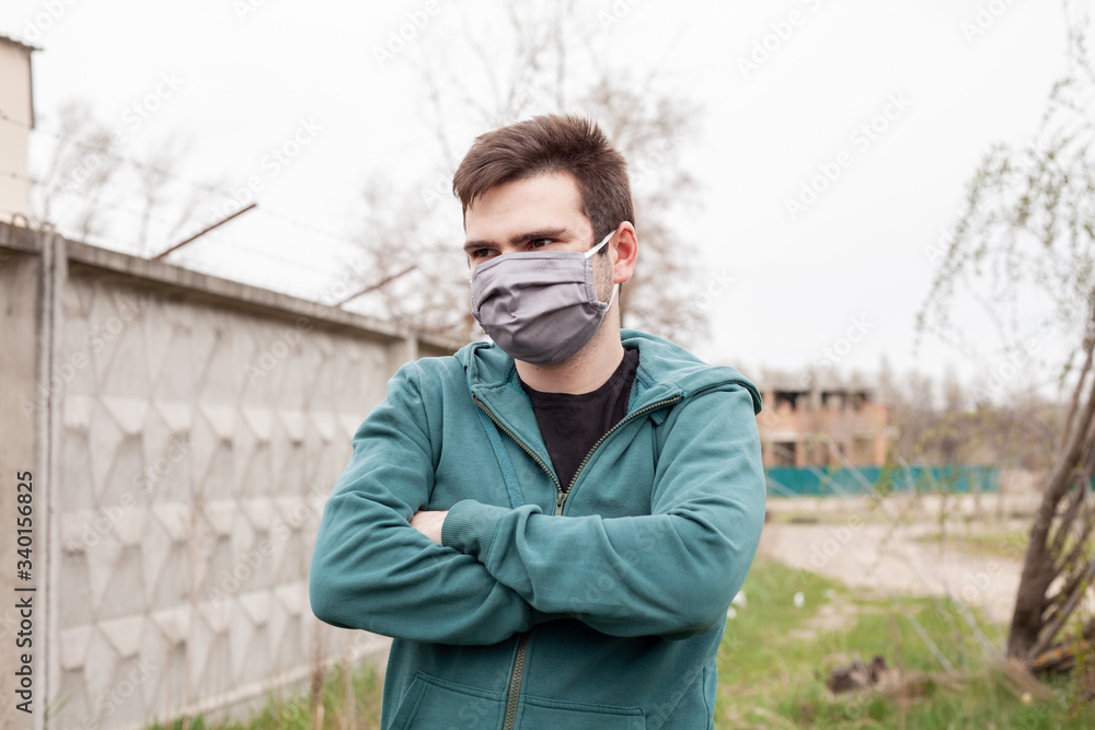 A man in a gray medical mask outdoors. His hands are folded. He is dressed in a green sweater. In the background is a gray, concrete fence, grass and trees. Quarantine time. A man in a gray medical mask outdoors. His hands are folded. He is dressed in a green sweater. In the background is a gray, concrete fence, grass and trees. Quarantine time.
