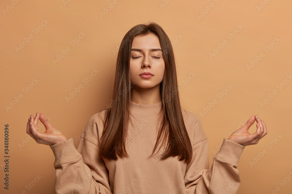 Peaceful calm relaxed young woman meditates indoor, keeps hands in zen gesture, closes eyes, practices yoga, finds peace inside of soul, reaches nirvana, dressed casually, poses indoor over beige wall Peaceful calm relaxed young woman meditates indoor, keeps hands in zen gesture, closes eyes, practices yoga, finds peace inside of soul, reaches nirvana, dressed casually, poses indoor over beige wall
