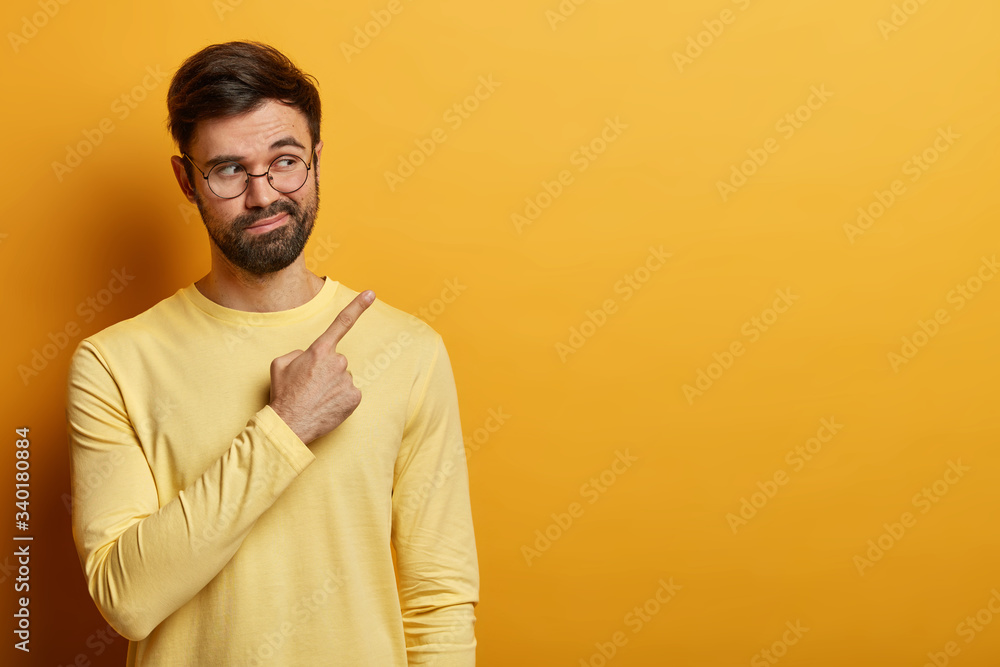 Curious bearded man points ay copy space on yellow background, demonstrates important information, found solution or answer on question, says his suggestion, wears yellow jumper, round spectacles Curious bearded man points ay copy space on yellow background, demonstrates important information, found solution or answer on question, says his suggestion, wears yellow jumper, round spectacles