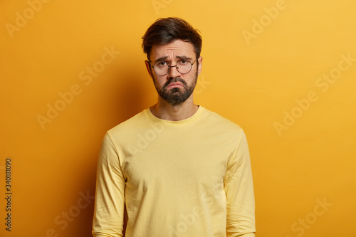 Troubled displeased bearded man frowns face, feels sad, distressed and upset, being bored sitting on quarantine, unhappy to miss good chance, dressed casually, isolated over yellow background.