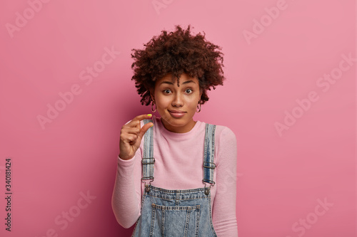 Photo of confused dark skinned woman shows small size with fingers, tells about decreased price, demonstrates tiny measure, asks for little offer, has disappointed expression, stands indoor.