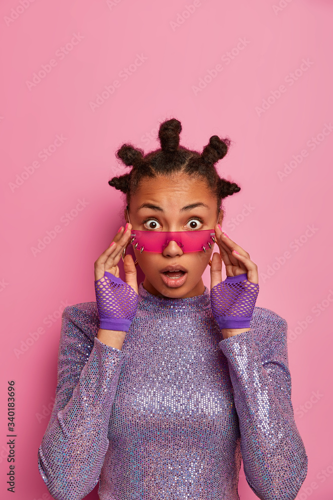 Astounded dark skinned hipster girl reacts on shocking relevation, gazes surprisingly through trendy rosy sunglasses, has hair buns, stands impressed indoor against pink pastel studio background Astounded dark skinned hipster girl reacts on shocking relevation, gazes surprisingly through trendy rosy sunglasses, has hair buns, stands impressed indoor against pink pastel studio background