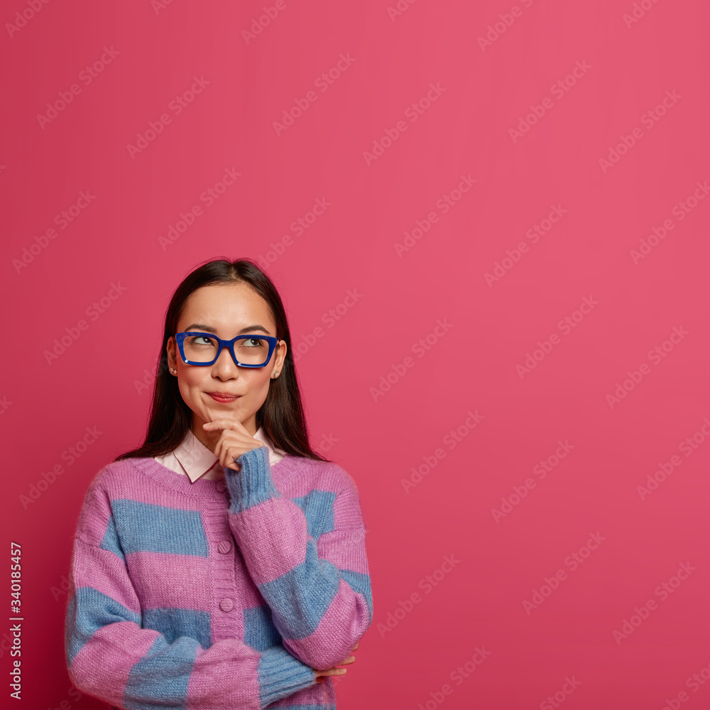Indecisive pretty schoolgirl holds chin and looks up thoughtful, makes choice with serious expression, imagines plan, thinks about something important, wears big transparent glasses, warm jumper Indecisive pretty schoolgirl holds chin and looks up thoughtful, makes choice with serious expression, imagines plan, thinks about something important, wears big transparent glasses, warm jumper