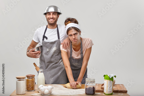 Fototapeta Naklejka Na Ścianę i Meble -  Tired young woman kneads dough for making homemade bread, happy husband in apron smiles happily and helps wife, has bowl on head, pose at kitchen near table with ingredients, try new recipe.