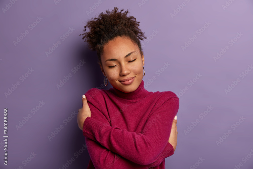 I love myself. Tender romantic woman embraces own body, hugs herself, closes eyes from pleasure, wears soft turtleneck just for cold weather, feels comfort, stands indoor against purple wall I love myself. Tender romantic woman embraces own body, hugs herself, closes eyes from pleasure, wears soft turtleneck just for cold weather, feels comfort, stands indoor against purple wall