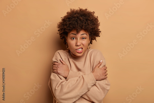 Displeased dark skinned woman hugs herself, feels freezed during cold day, winter is coming, clenches teeth, shakes during chilling weather, stands indoor. Fearful female afraids of something © wayhome.studio 