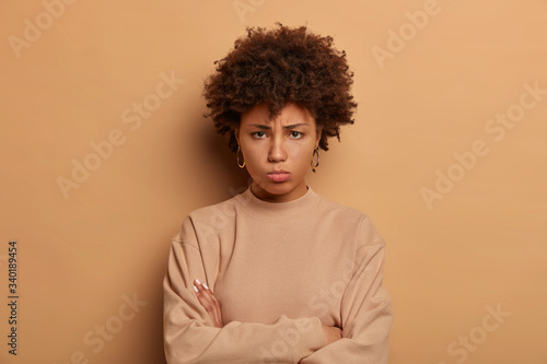 Photo of troubled sad woman stands upset and offended, keeps arms folded, looks with regret from under forehead, wears casual beige jumper, expresses negative emotions, blows cheeks from anger