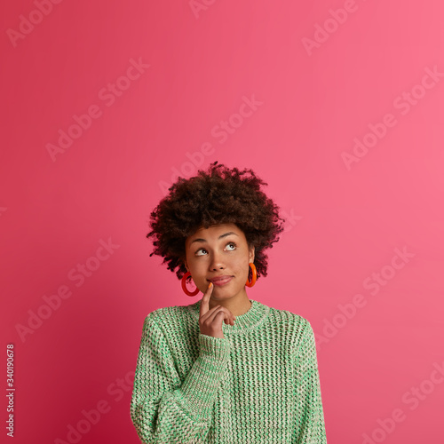 Curious African American woman concentrated above  tries to decide something  stands in thoughtful pose  keeps finger near lips  wears knitted sweater  isolated on pink wall  blank space upwards