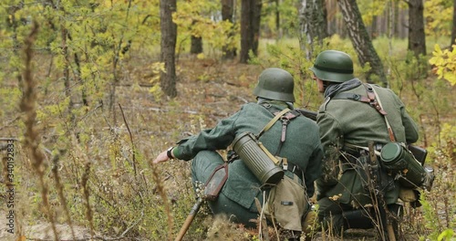 Two Re-enactors Dressed As German Wehrmacht Infantry Soldiers In World War II Soldier Sitting In Ambush In Autumn Forest And Looking At Side. Historical Re-enactment. 4K photo