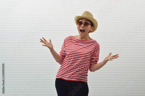 Surprised young asian woman wearing red stripped shirt, sunglasses and straw hat standing over white wall background, Exciting facial expression, Business summer holiday concept