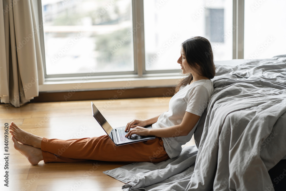 Beautiful young brunette girl working on a laptop, sitting on the floor near the bed by the panoramic window with a beautiful view from the high floor. Stylish modern interior. A cozy workplace Beautiful young brunette girl working on a laptop, sitting on the floor near the bed by the panoramic window with a beautiful view from the high floor. Stylish modern interior. A cozy workplace