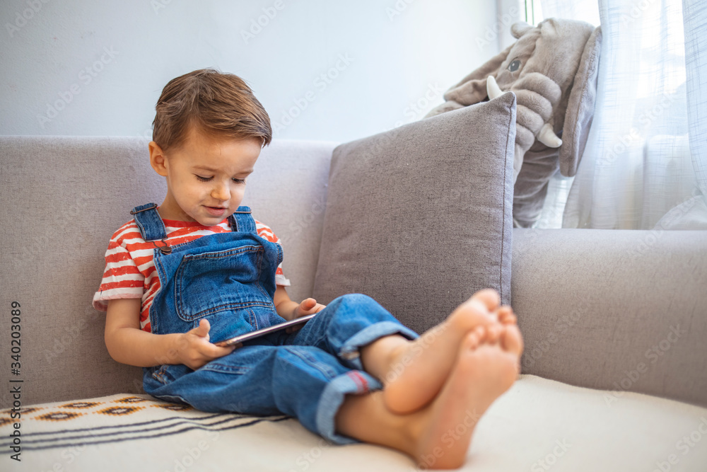 Cute boy using a mobile phone. A little boy looking to smartphone lying on the couch at home. Toddler boy using tablet or smartphone. Cute three years old boy sitting at home using digital device. Cute boy using a mobile phone. A little boy looking to smartphone lying on the couch at home. Toddler boy using tablet or smartphone. Cute three years old boy sitting at home using digital device.