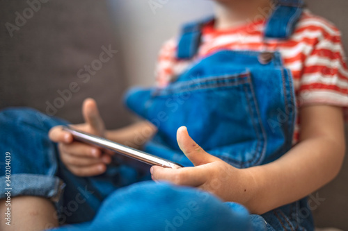 Little boy using smart phone in the living room. Education and learning concept. Boy sits on sofa plays with smartphone. Child and electronic devices concept. Portrait of toddler with smartphone