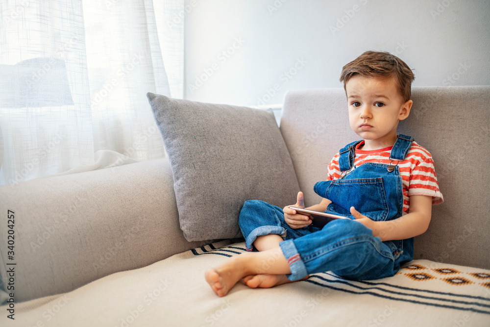 Adorable toddler boy sitting on the sofa in the living room and playing with smartphone. Child learning how to use smartphone. Boy texting on the phone. - technology and lifestyle concept. Adorable toddler boy sitting on the sofa in the living room and playing with smartphone. Child learning how to use smartphone. Boy texting on the phone. - technology and lifestyle concept.