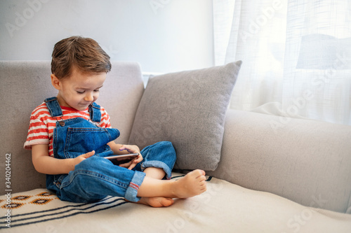 Cute boy using a mobile phone. A little boy looking to smartphone lying on the couch at home. Toddler boy using tablet or smartphone. Cute three years old boy sitting at home using digital device.