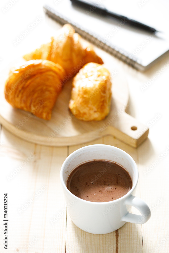 A cup of hot chocolate and croissant on wooden background with notebook. Work from home. A cup of hot chocolate and croissant on wooden background with notebook. Work from home.