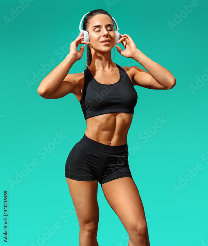 Sporty girl listening music with her headphones. Photo of attractive girl in black sportswear on turquoise background. Full length © Romario Ien