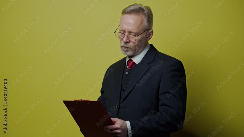 Senior man in costume is writing. Mature businessman in glasses holding a folder and thinking. Portrait of an old professional entrepreneur on yellow background. Senior man in costume is writing. Mature businessman in glasses holding a folder and thinking. Portrait of an old professional entrepreneur on yellow background.