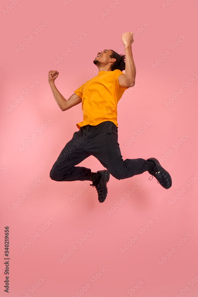Full length shot of a cheerful funny black man in sneakers, crazy flying carefree jumping with raised arms, celebrating victory, having fun, rest, relax and leisure, isolated on pink background