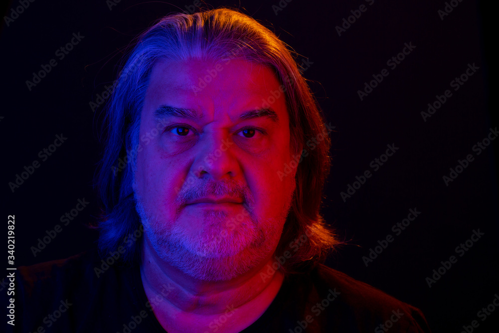 Closeup headshot of man. he is serious and looking to the camera. The studio portrait has lighting its with gel colored flashlights, red to her left, blue to her right and orange on top.