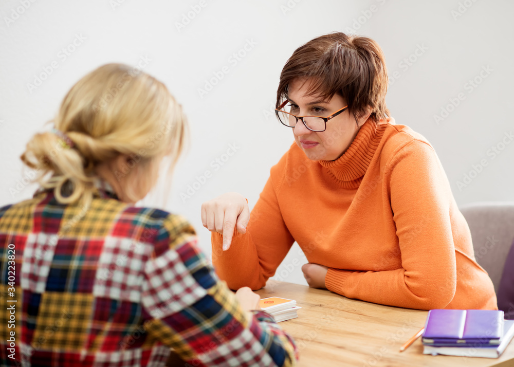 Two women talking each other. Woman wearing glasses, orange sweater listening carefully other woman wearing colorful jacket sitting in front of her in office Two women talking each other. Woman wearing glasses, orange sweater listening carefully other woman wearing colorful jacket sitting in front of her in office
