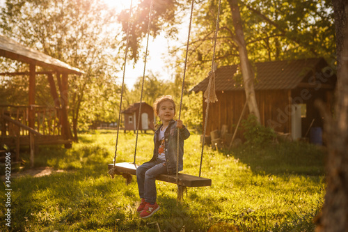 A little girl in a denim suit swings on a suspended swing to a tree. A girl in blue jeans on a swing with long ropes