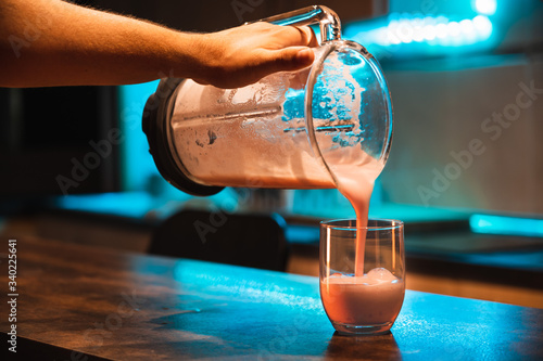 Close up of a hand pouring strawberry milkshake into a glass at a kitchen bar with orange ambient light and a blue teal light that comes from a light strip situated at the top of the kitchen surface