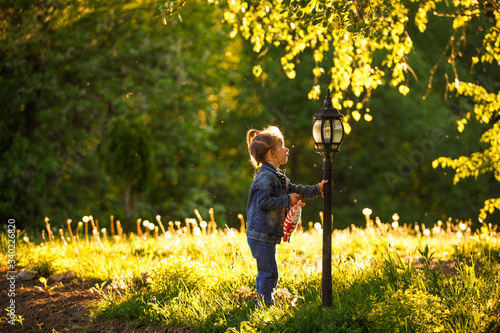 A little girl in a denim suit touches a lantern in the Park. Curious girl in the green grass in the summer sunlight
