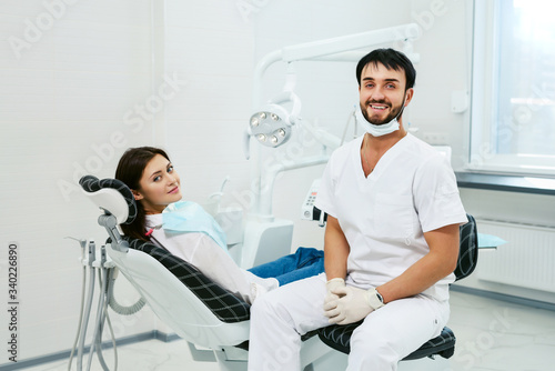 Portrait of a friendly smiling male dentist with patient in the office of a modern dental clinic. Doctor in disposable medical facial mask.