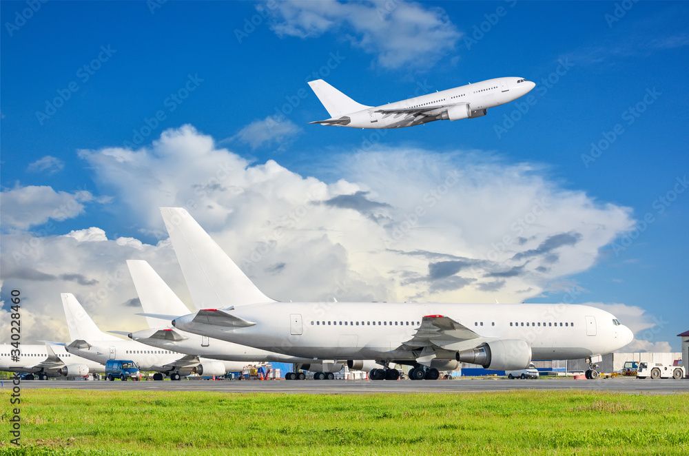 View of the standing aircraft at the airport and airplane taking off in cloudy sky. View of the standing aircraft at the airport and airplane taking off in cloudy sky.