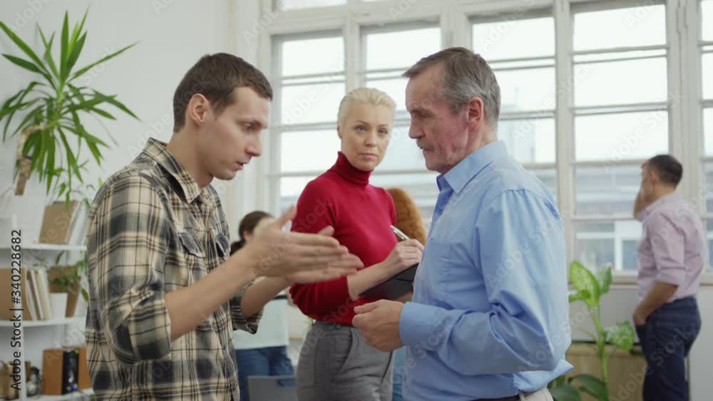Side view of young and senior men talking during mental health exercise at group therapy session, female therapist with clipboard walking in background and making notes Side view of young and senior men talking during mental health exercise at group therapy session, female therapist with clipboard walking in background and making notes