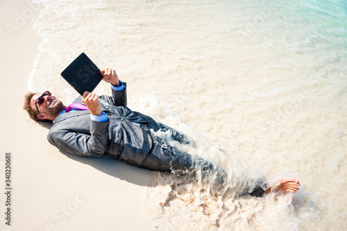 Barefoot businessman relaxing with a tablet computer on the shore of a tropical beach with waves breaking over his suit